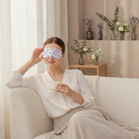 SlumberHush WarmEye steam mask with purple leaf pattern, worn by a woman relaxing on a sofa with a cup of tea.